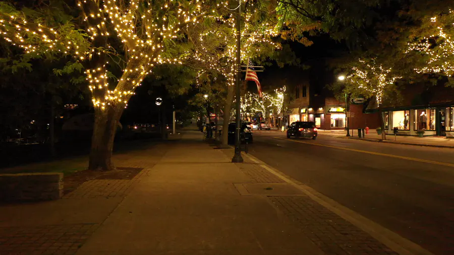 A wide downtown sidewalk at night beneath street trees wrapped in warm white lights, with storefronts glowing across the street.