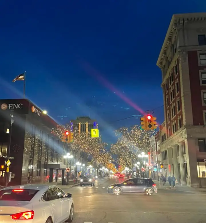 A downtown street at dusk lined with trees wrapped in warm white lights, with traffic signals, a crosswalk and historic brick buildings.