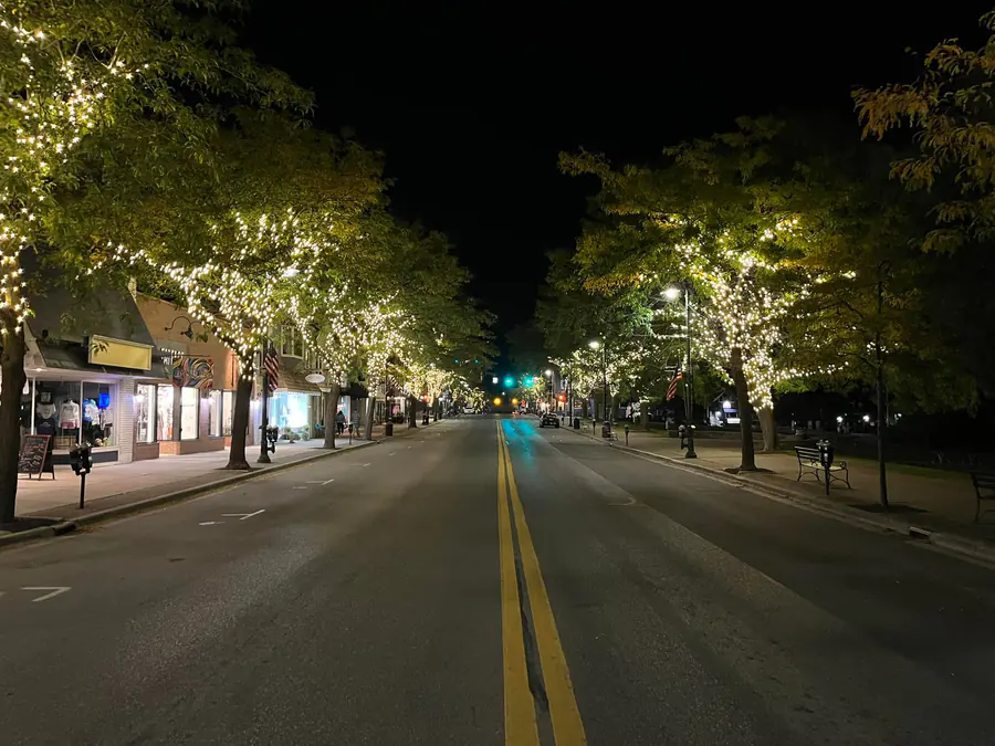 An empty downtown main street at night with trees on both sides wrapped in warm white lights above storefronts and sidewalks.