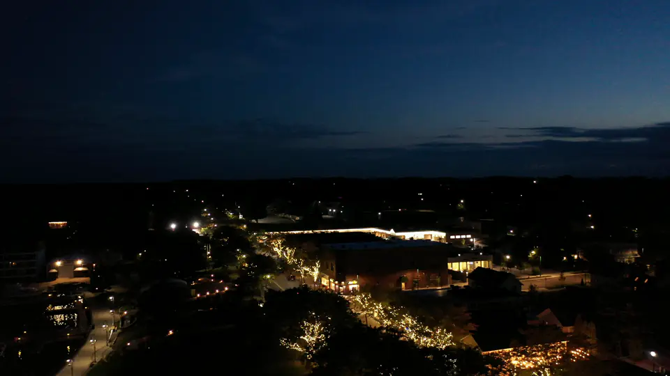 A slow aerial pan across a small downtown at blue hour, with warm white wrapped trees glowing under a deep blue sky.